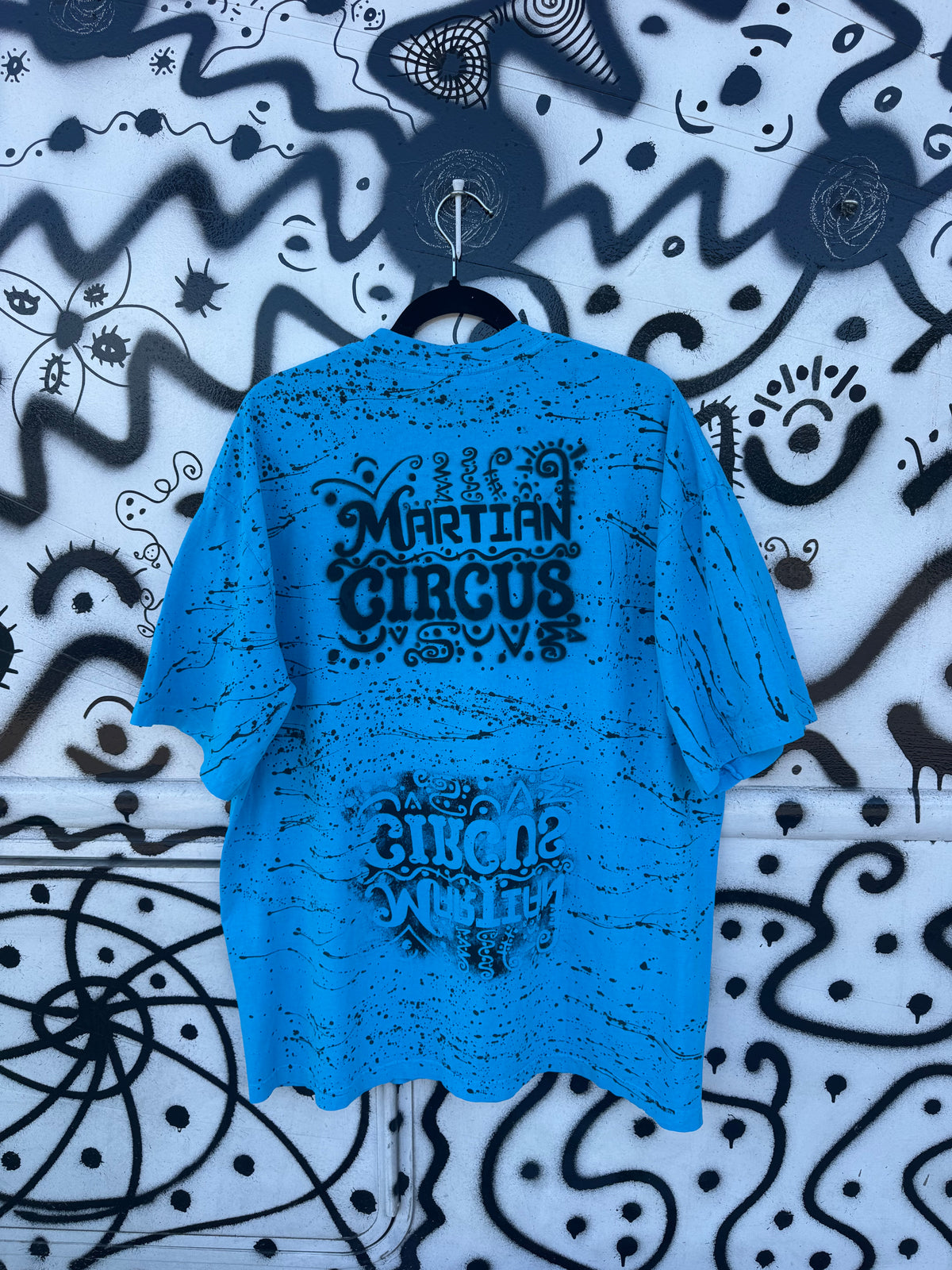 A bright blue t-shirt with black distressed graphics reading 'MARTIAN CIRCUS'. hanging on a black hanger against a black and white graffiti-patterned wall.