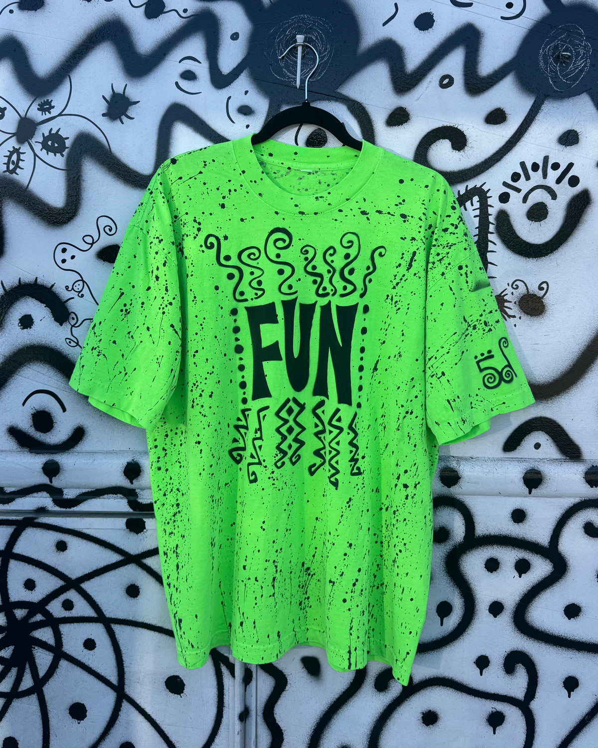 A bright neon green short-sleeve t-shirt with black distressed splatter details and a central graphic reading 'FUN' in black, hanging on a black hanger against a white wall covered in black graffiti-style drawings.