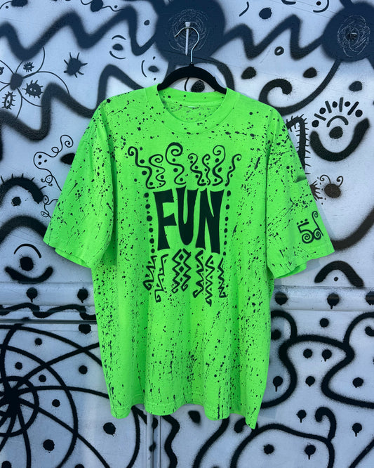A bright neon green short-sleeve t-shirt with black distressed splatter details and a central graphic reading 'FUN' in black, hanging on a black hanger against a white wall covered in black graffiti-style drawings.