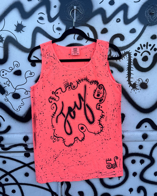 A bright coral pink sleeveless tank top with a black distressed graphic reading 'joy' in script, hanging on a black hanger against a blue wall covered in black graffiti-style drawings.