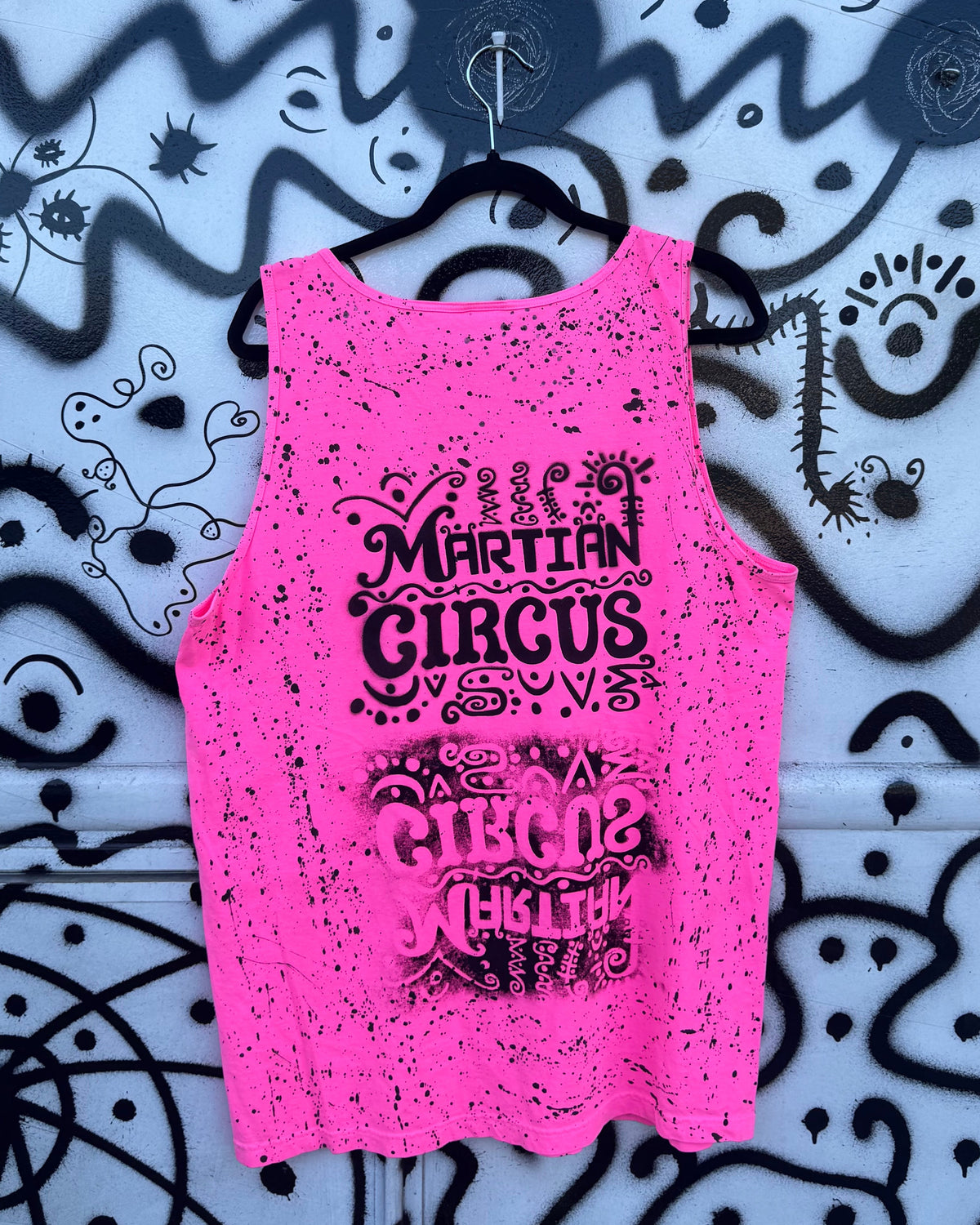 A bright pink sleeveless tank top with black graphic text reading 'MARTIAN CIRCUS' hanging on a black hanger against a blue and black abstract patterned background.